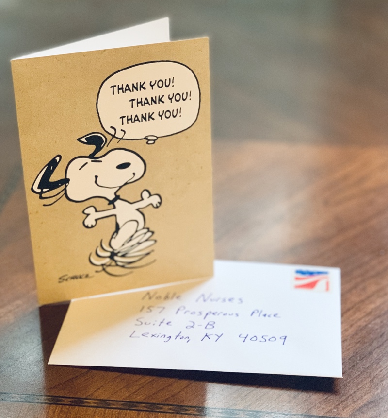 A photo of a greeting card on a desk. The card is light brown with Snoopy dancing and saying Thank You three times. The white envelope is addressed to Noble Nurses, LLC.